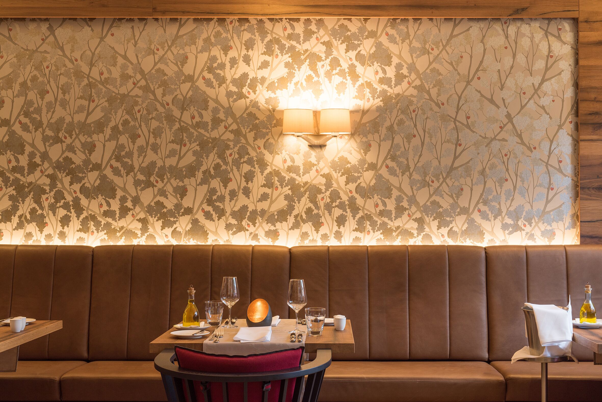Ein Restaurantinneres mit einer Essecke mit Sitzelementen, einem Tisch für zwei Personen gedeckt mit Weingläsern und einer Weinflasche, und einer Wand mit floralem Muster.