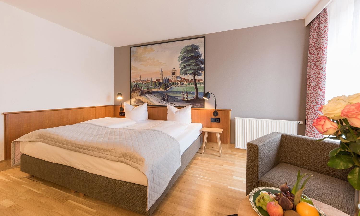 A hotel room with a large bed, a wooden headboard, a nightstand with a lamp, a painting on the wall, and a small table with fruit and flowers.