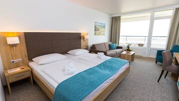 Ein Hotelzimmer mit einem großen Bett, einer Sitzecke mit Sessel und Tisch, einem kleinen Schreibtisch und einem Fenster mit Meerblick.