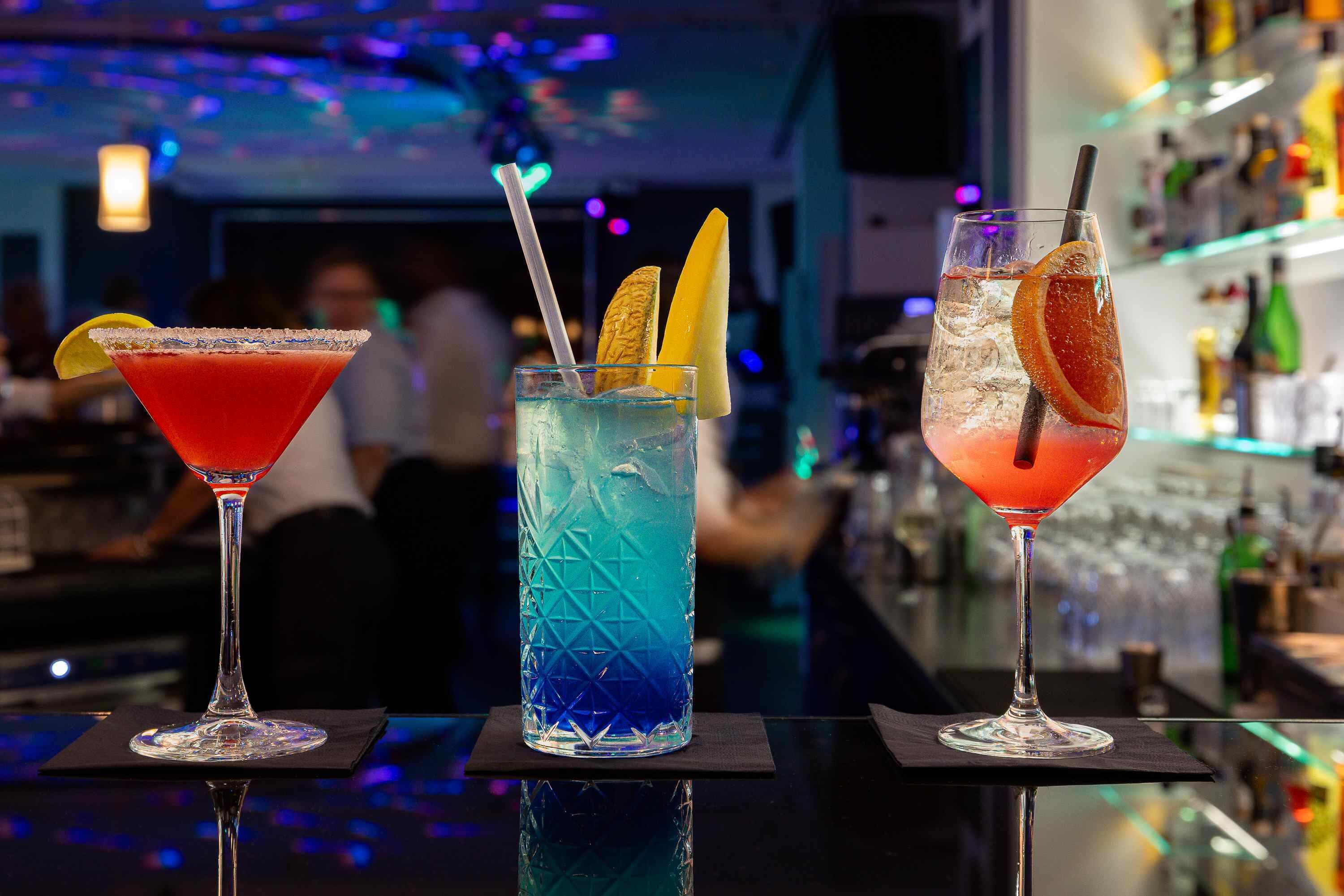 Tres cócteles sobre una barra, con un fondo desenfocado de un bar y gente.