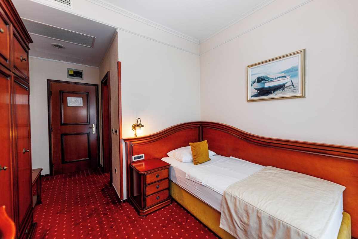 A hotel room with a wooden bed, a nightstand, a red carpeted floor, a door with a sign, and a framed picture on the wall.