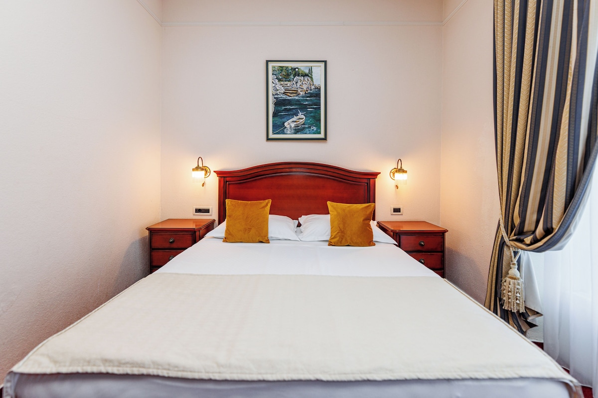 Una cama bien hecha con cabecera, dos mesitas de noche con lámparas, un cuadro en la pared y una ventana con cortinas.