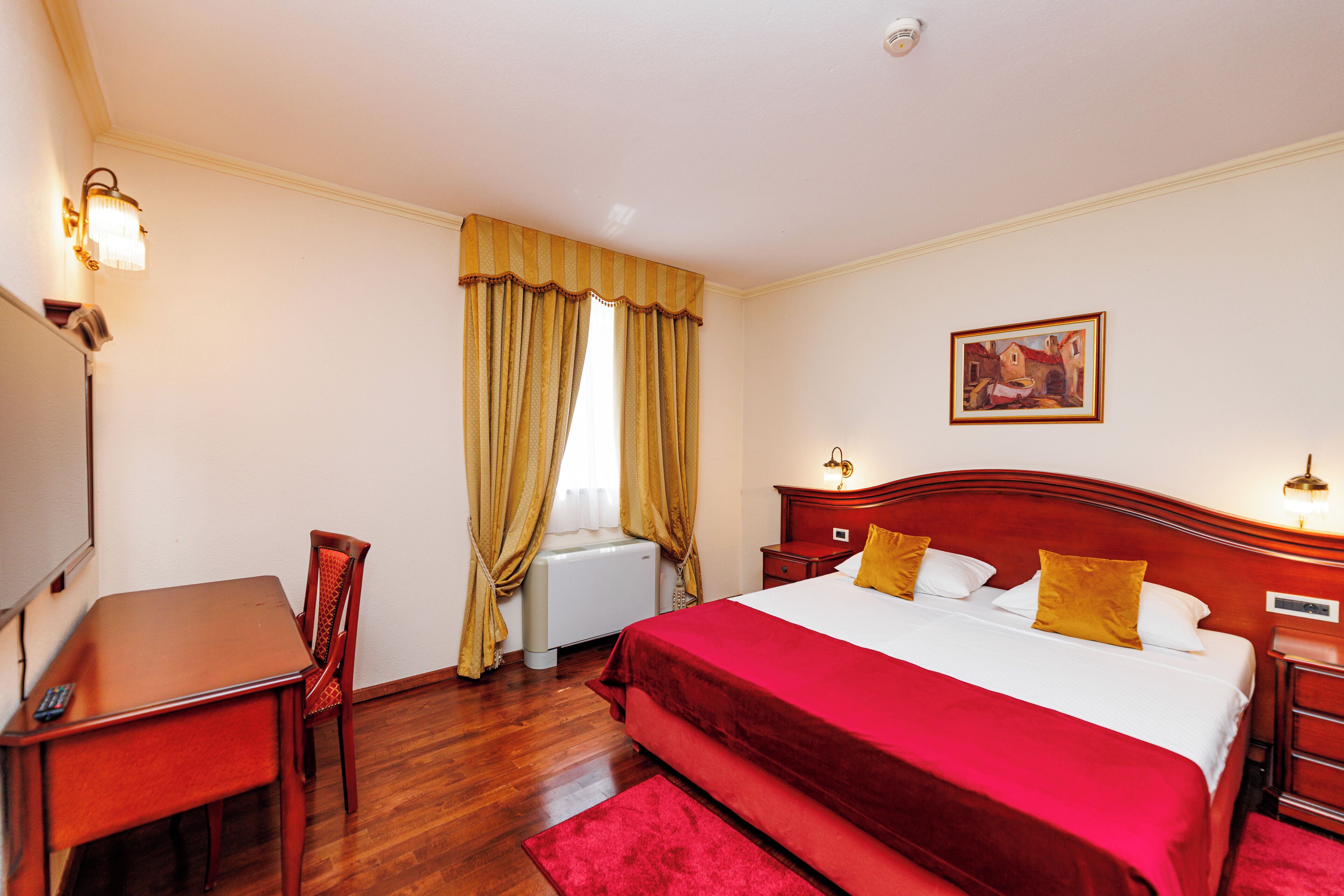 A hotel room with a wooden bed, a desk with a chair, a radiator, and a painting on the wall.