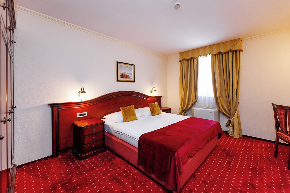 A hotel room with a large bed, wooden headboard, two bedside tables with lamps, a red carpet, and a window with curtains.