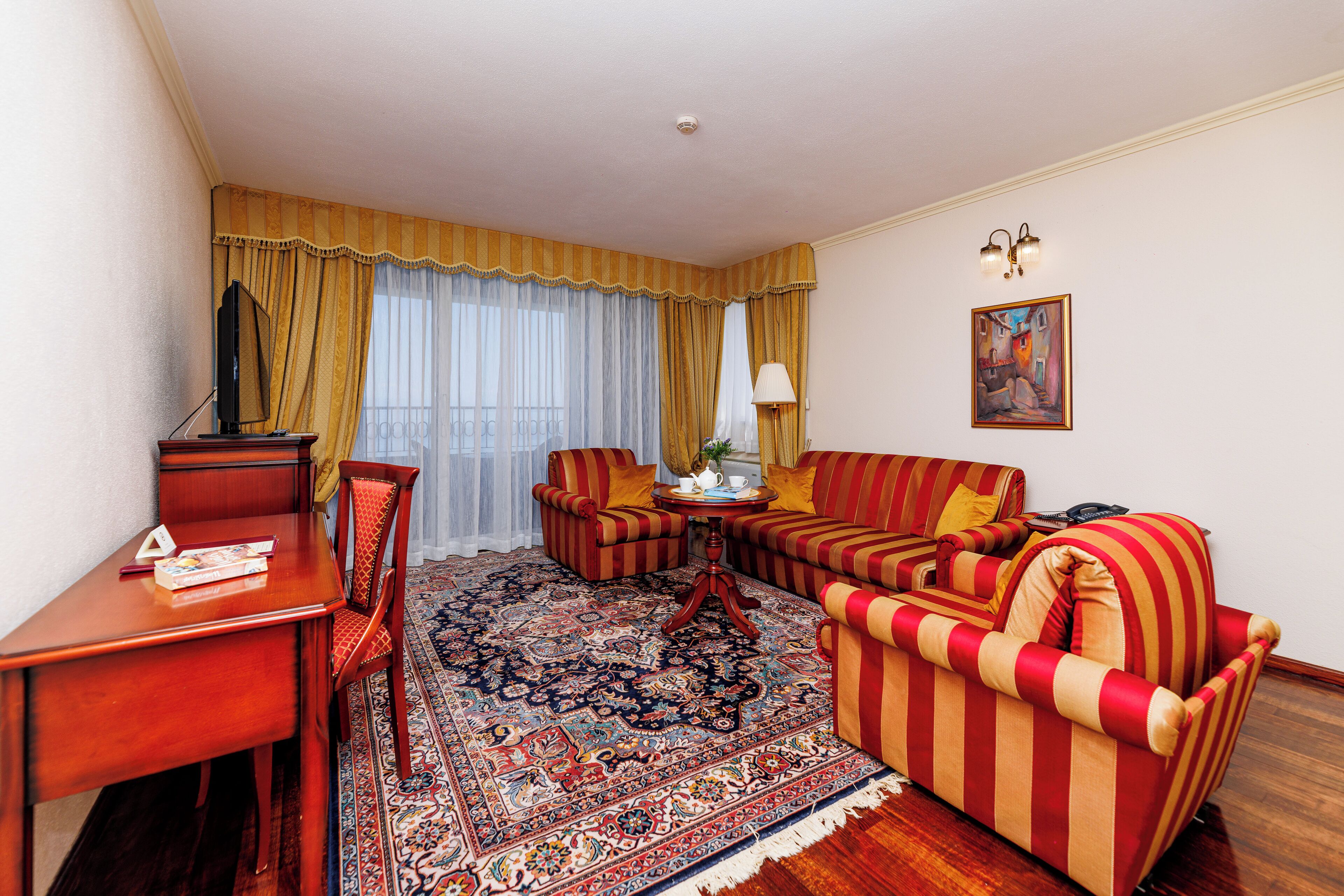A hotel room with a wooden desk, striped armchairs, a sofa, a patterned rug, a painting on the wall, and a window with curtains.