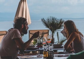 A couple dining at an outdoor table with a view of the sea, surrounded by potted plants and a beach umbrella.