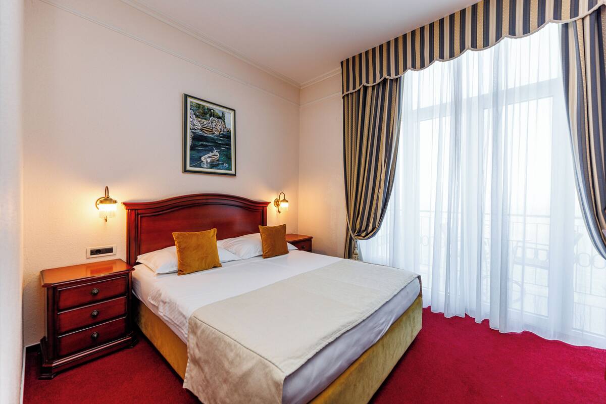 A hotel room with a large bed, two bedside tables with lamps, a headboard, a painting on the wall, and a window with curtains.