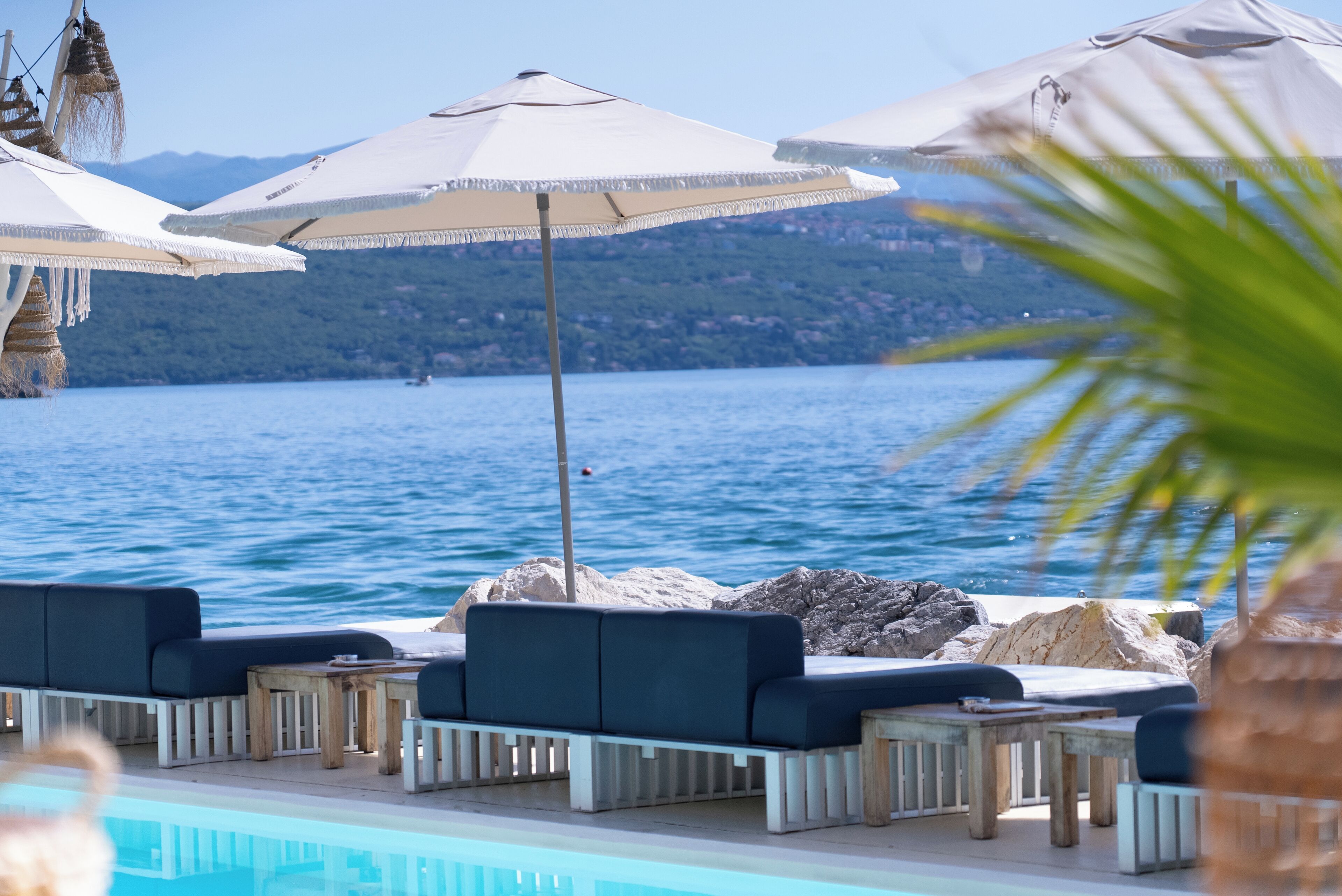 A poolside area with lounge chairs and umbrellas, overlooking a body of water and a mountainous landscape.