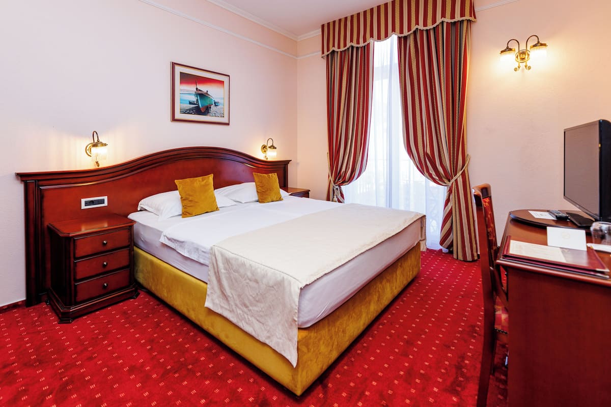 A hotel room with a large bed, a desk, a chair, a nightstand, a lamp, a painting on the wall, and a window with curtains.