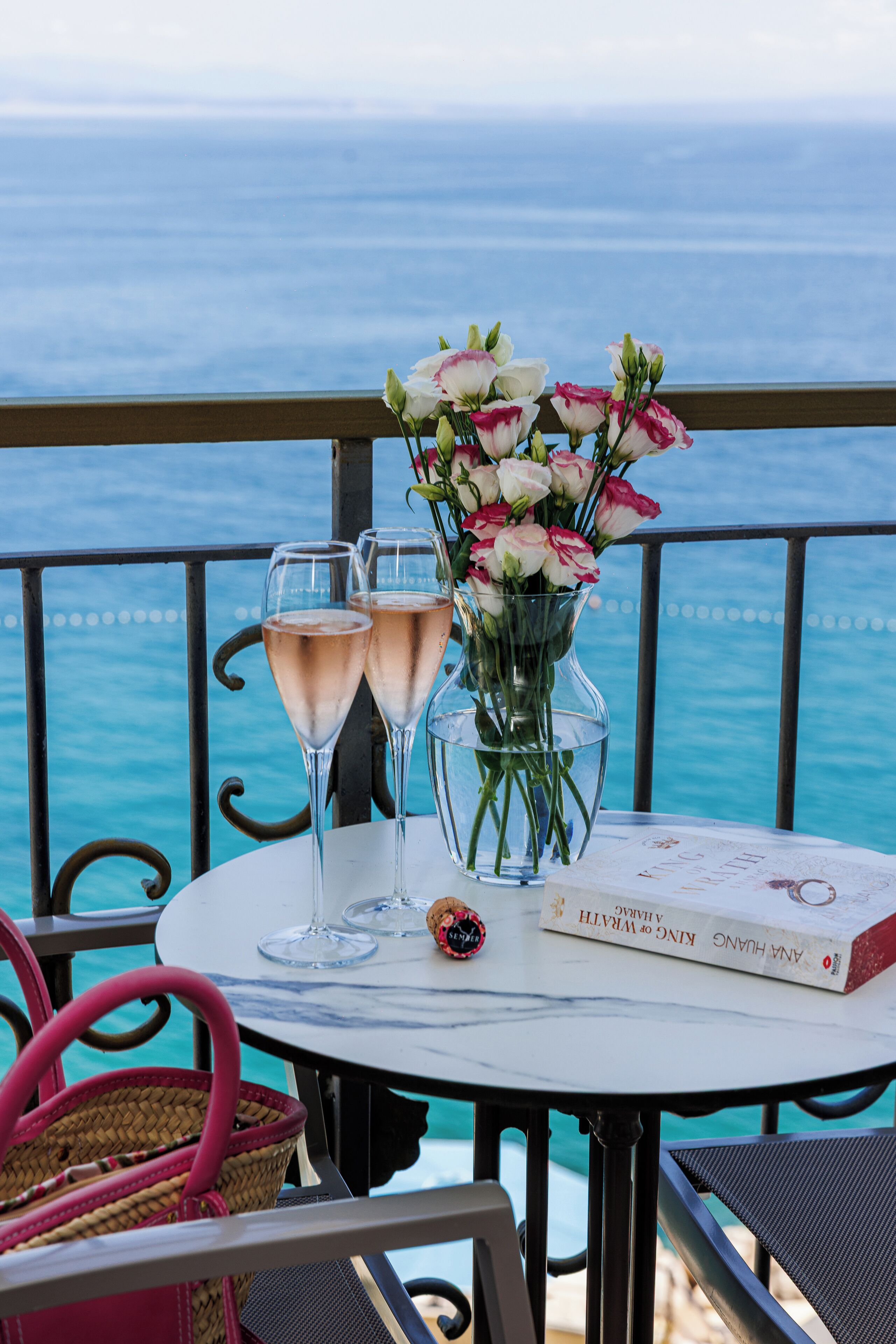 A table with two champagne glasses, a vase of flowers, and a book titled "King & I" on a balcony overlooking the sea.