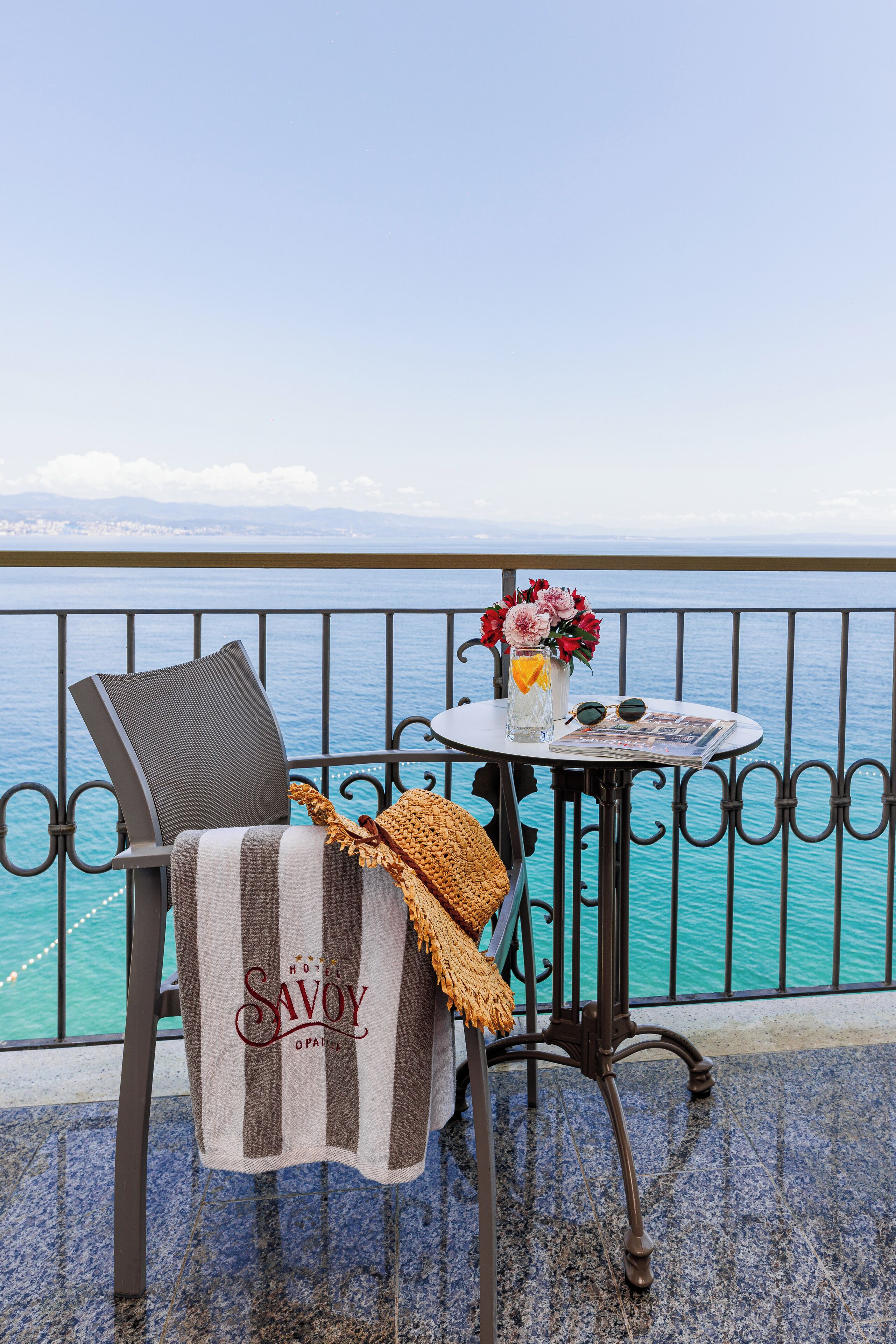 Ein Balkon mit einem Stuhl, einem kleinen Tisch und Meerblick, dazu ein Handtuch mit dem Schriftzug 'HOTEL SAVOY'.