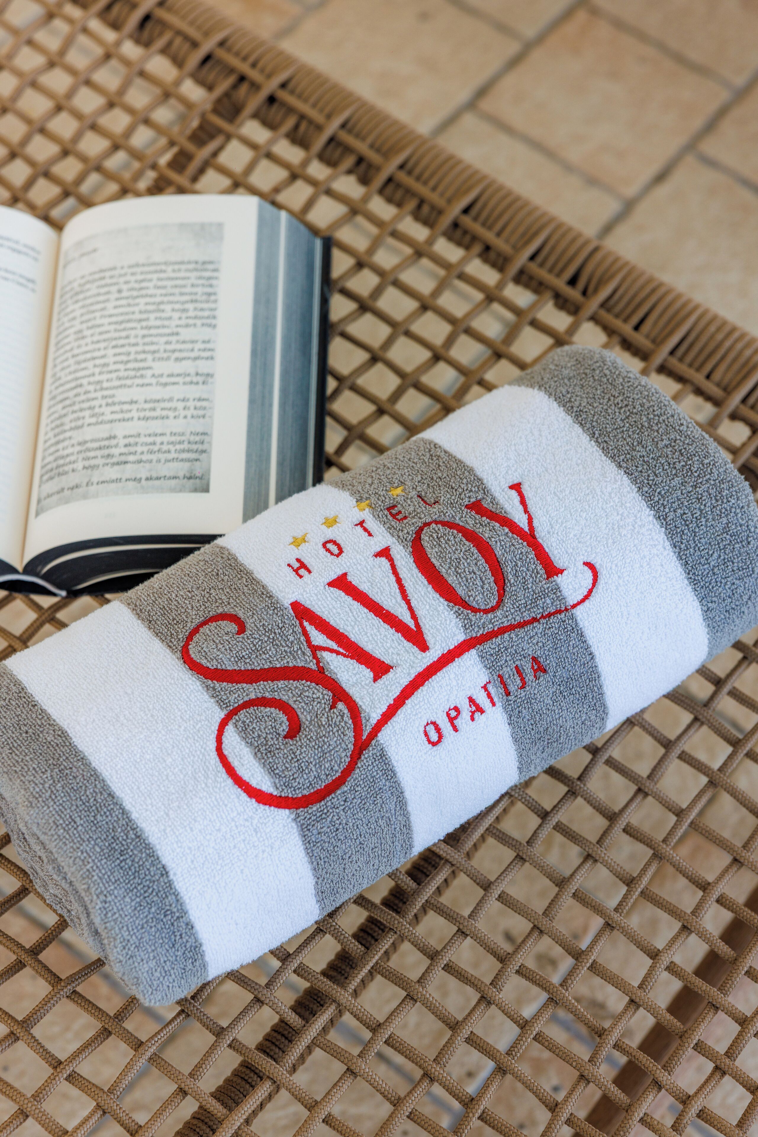 Une serviette sur laquelle est brodé le nom « HÔTEL SAVOY », posée sur une surface tissée à côté d’un livre ouvert.