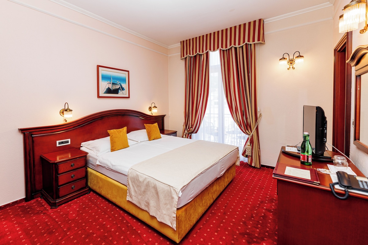 Habitación de hotel con una cama grande, una mesita de noche con lámpara, un escritorio con computadora y una ventana con cortinas.