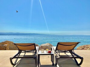 Dos sillones de relax mirando al mar, con una mesa entre ellos, una bebida en un vaso y una taza sobre la mesa.