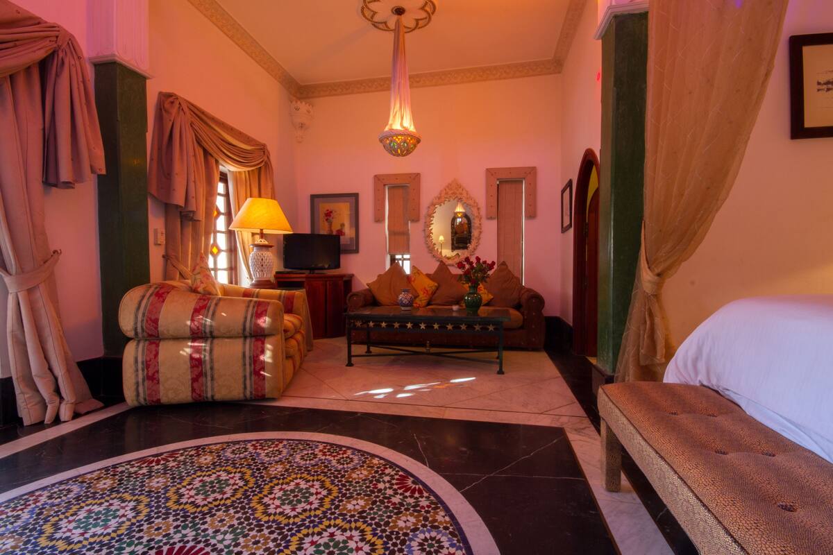 A hotel room with a sofa, a small table with a lamp, a television, a decorative mirror, and a bed.