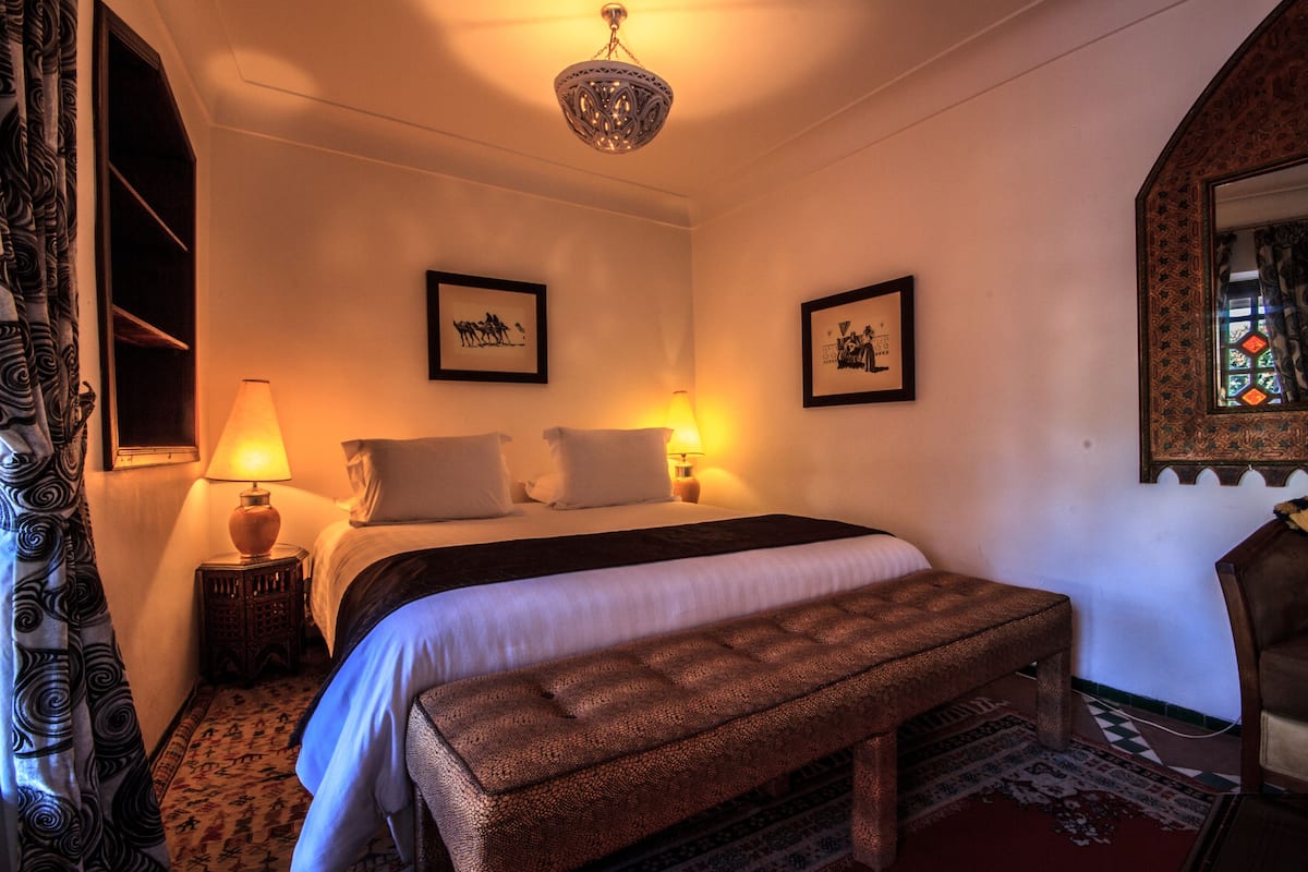A bedroom with a bed, two bedside lamps, a bench, a patterned rug, and two framed pictures on the wall.