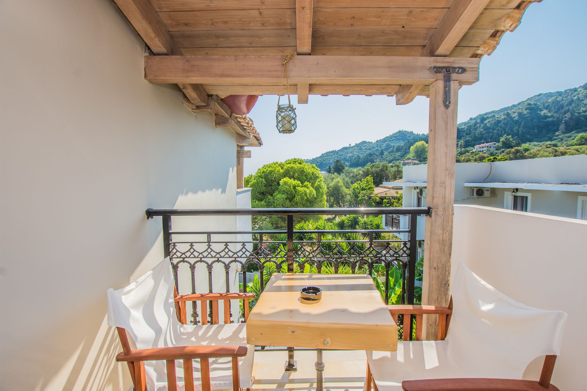Un balcone con tavolo e sedie in legno, ringhiera in metallo e vista su alberi verdi e edifici sullo sfondo.