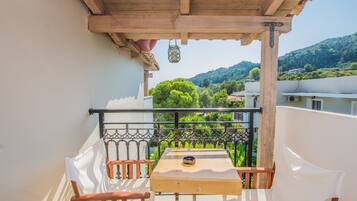Un balcone con tavolo e sedie in legno, ringhiera in metallo e vista su alberi verdi e edifici sullo sfondo.