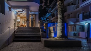 The entrance of the HNIXOXE INIOCHOS HOTEL with a staircase leading to the lobby, a large planter with a tree, and a sign above the entrance.