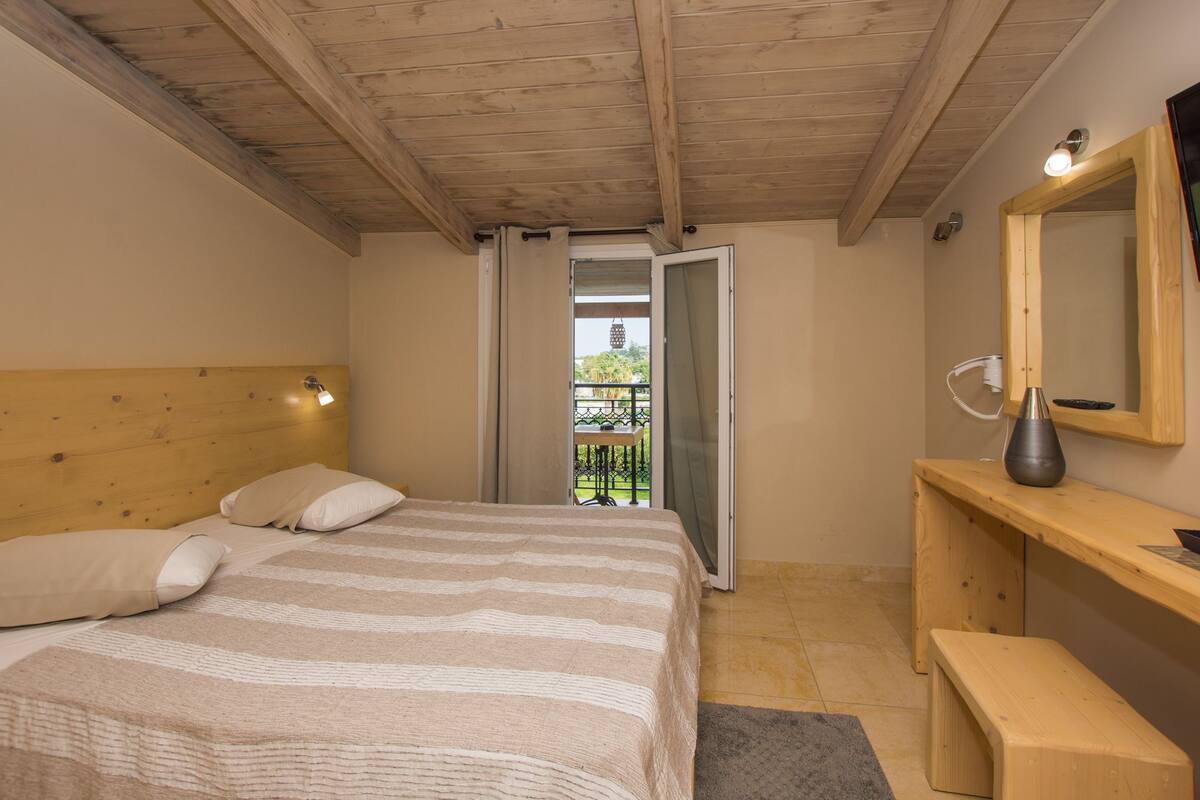 A bedroom with a wooden ceiling, a bed with striped bedding, a bedside table, a mirror, and a view of a balcony.