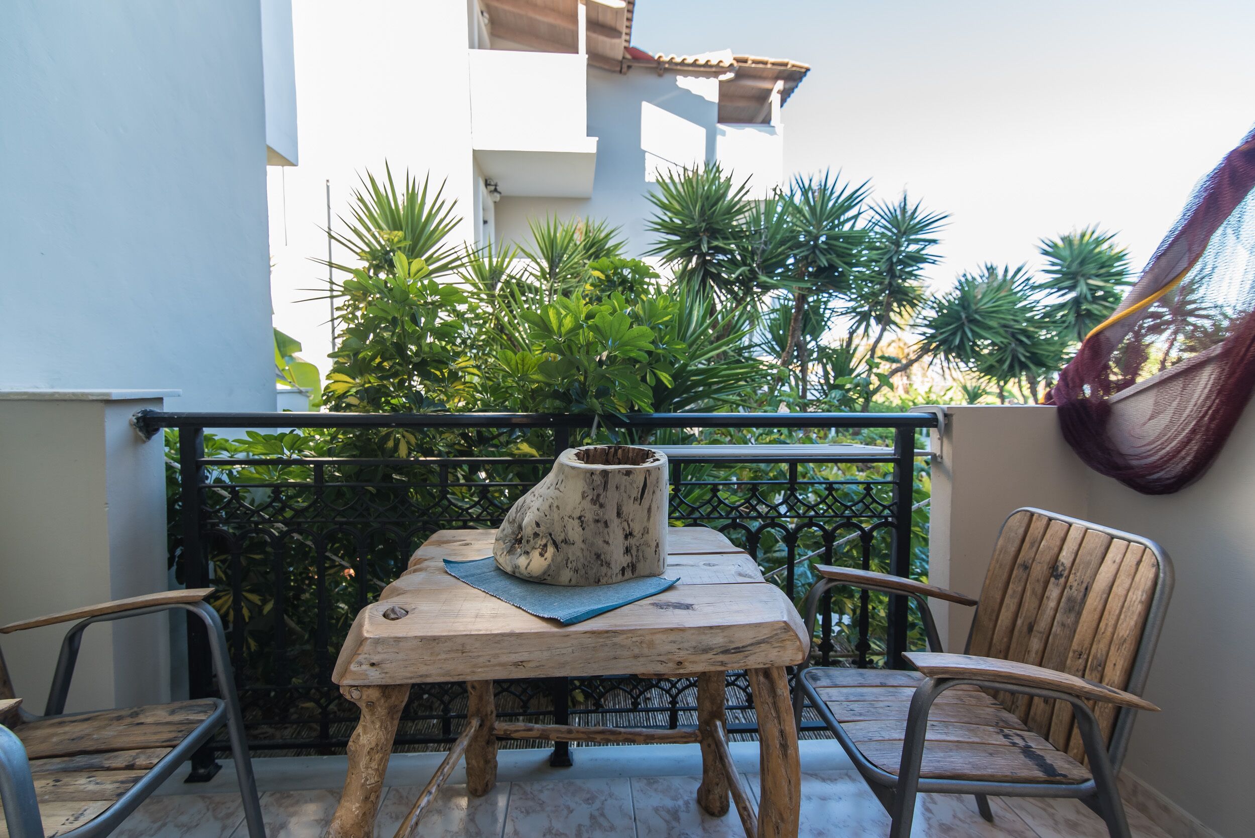 Een houten tafel met een vaas en een blauwe mat op een balkon met metalen stoelen, omgeven door groen en een wit gebouw.