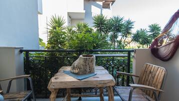Een houten tafel met een vaas en een blauwe mat op een balkon met metalen stoelen, omgeven door groen en een wit gebouw.