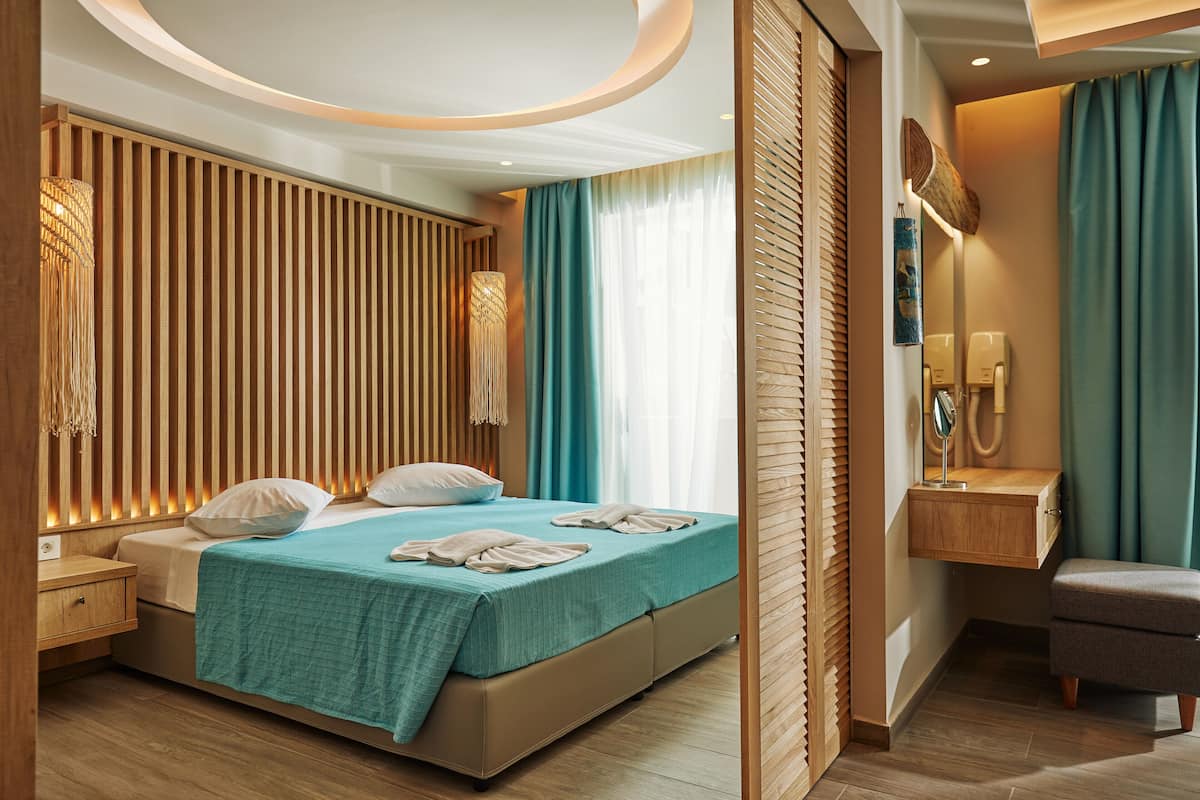 A modern hotel room with a large bed, a wooden headboard, a bedside table, a nightstand, a small seating area, and a bathroom with a mirror and towel rack.