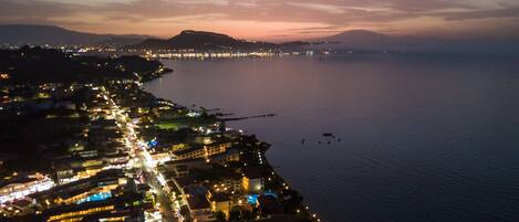 An aerial view of a coastal city at dusk with illuminated buildings along the waterfront and a calm sea.
