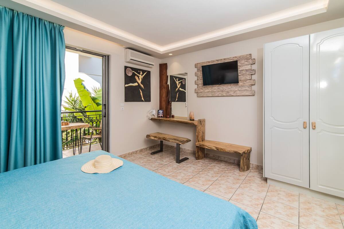 A bedroom with a bed, a TV mounted on a wooden wall, a wardrobe, and a balcony with a table and chairs.