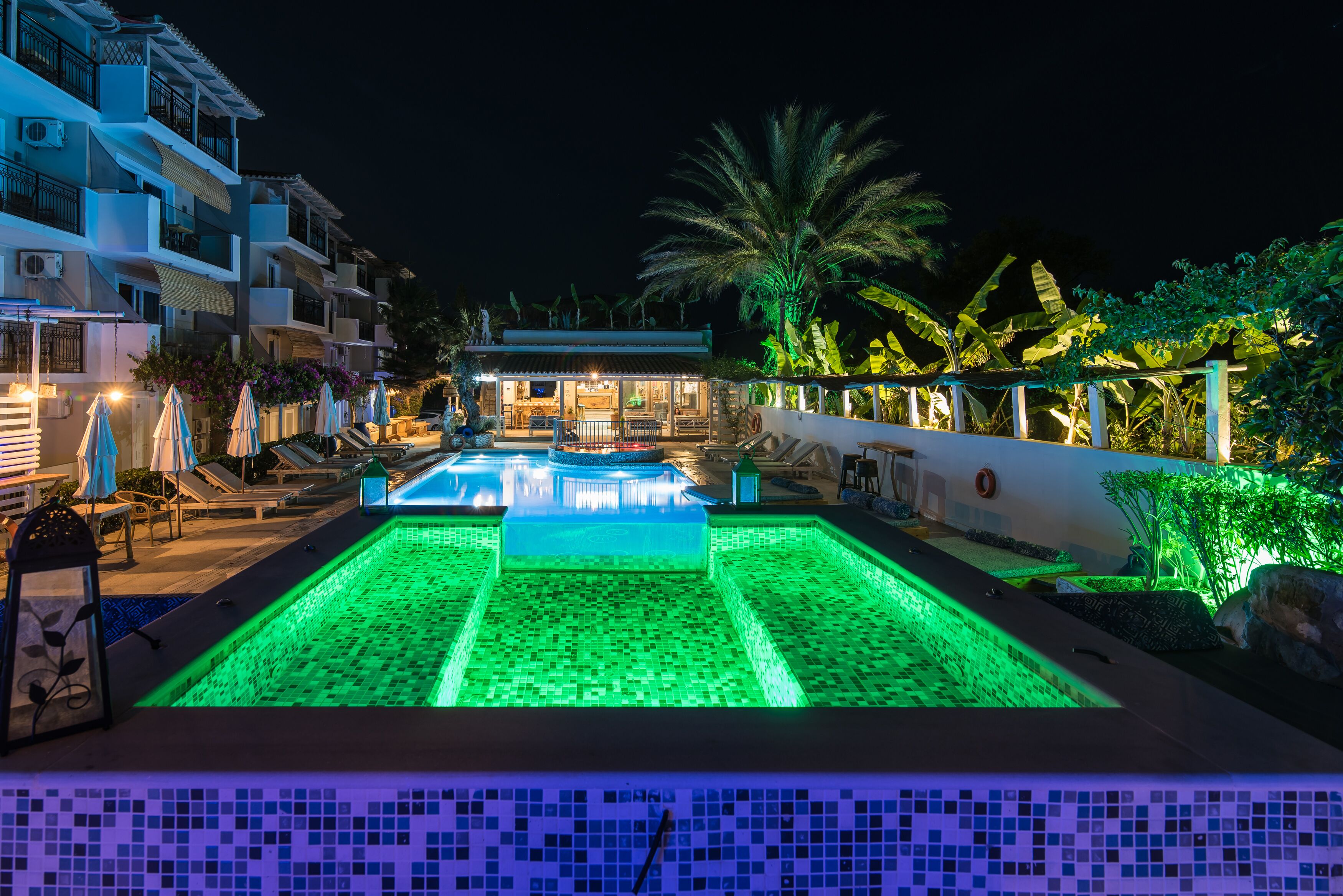 A swimming pool with green lighting, surrounded by lounge chairs and umbrellas, with a building and palm trees in the background.