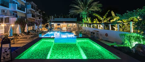 A swimming pool with green lighting, surrounded by lounge chairs and umbrellas, with a building and palm trees in the background.