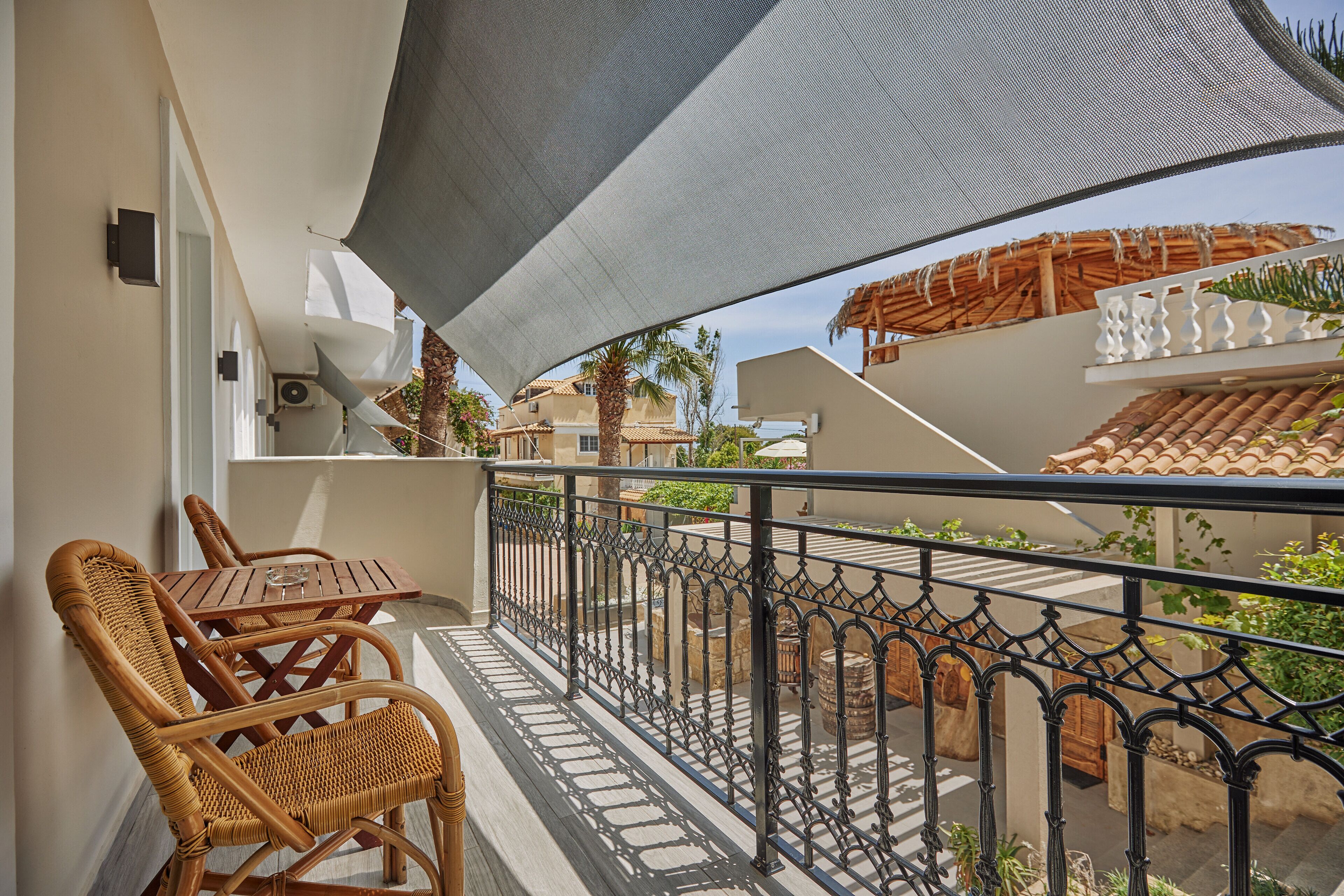 A balcony with wicker chairs, a table, and a view of a courtyard with plants and a building with a tiled roof.