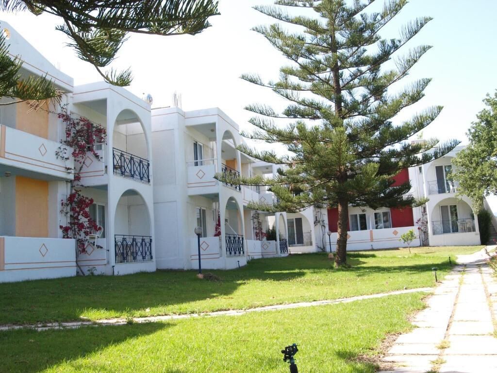 Una hilera de edificios blancos con balcones, un césped cuidado y un gran pino en primer plano.