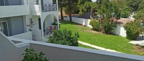Un balcone con una pianta in vaso, un tavolo bianco e vista su un cortile con alberi e una piscina.