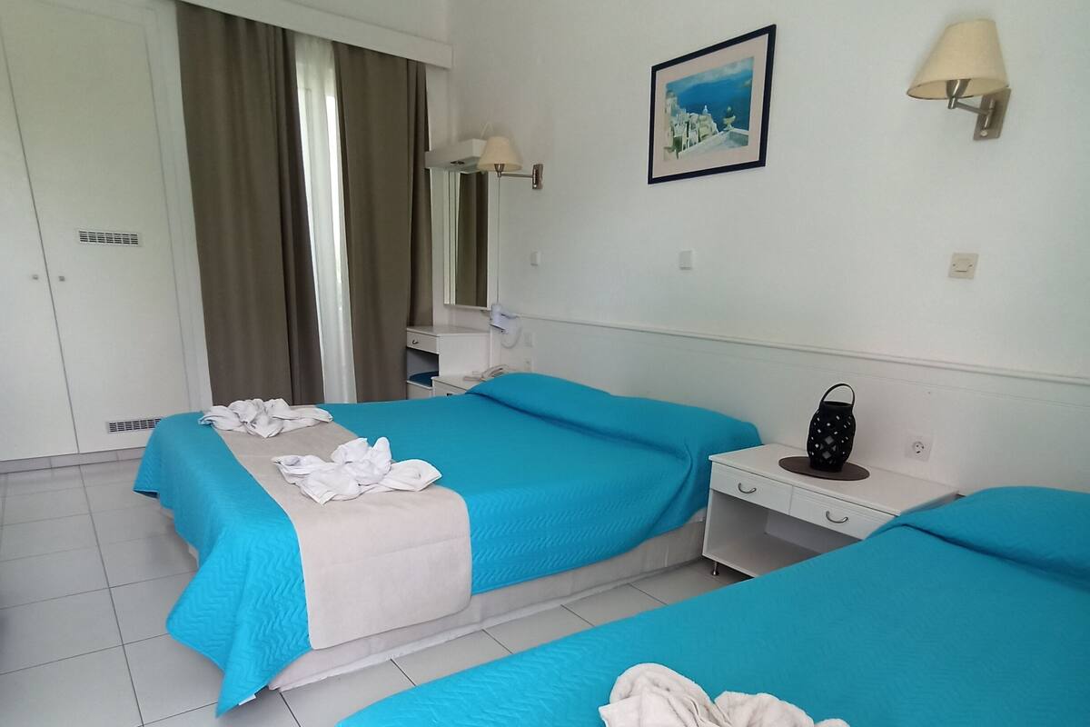 Hotellihuone, jossa on kaksi sänkyä. Kummassakin sängyssä on sininen peitto ja valkoiset pyyhkeet on aseteltu kukka-aiheiseksi koristeeksi. Huoneessa on yöpöytä, jossa on lamppu, sekä pieni pöytä, jonka päällä on koriste-esine.