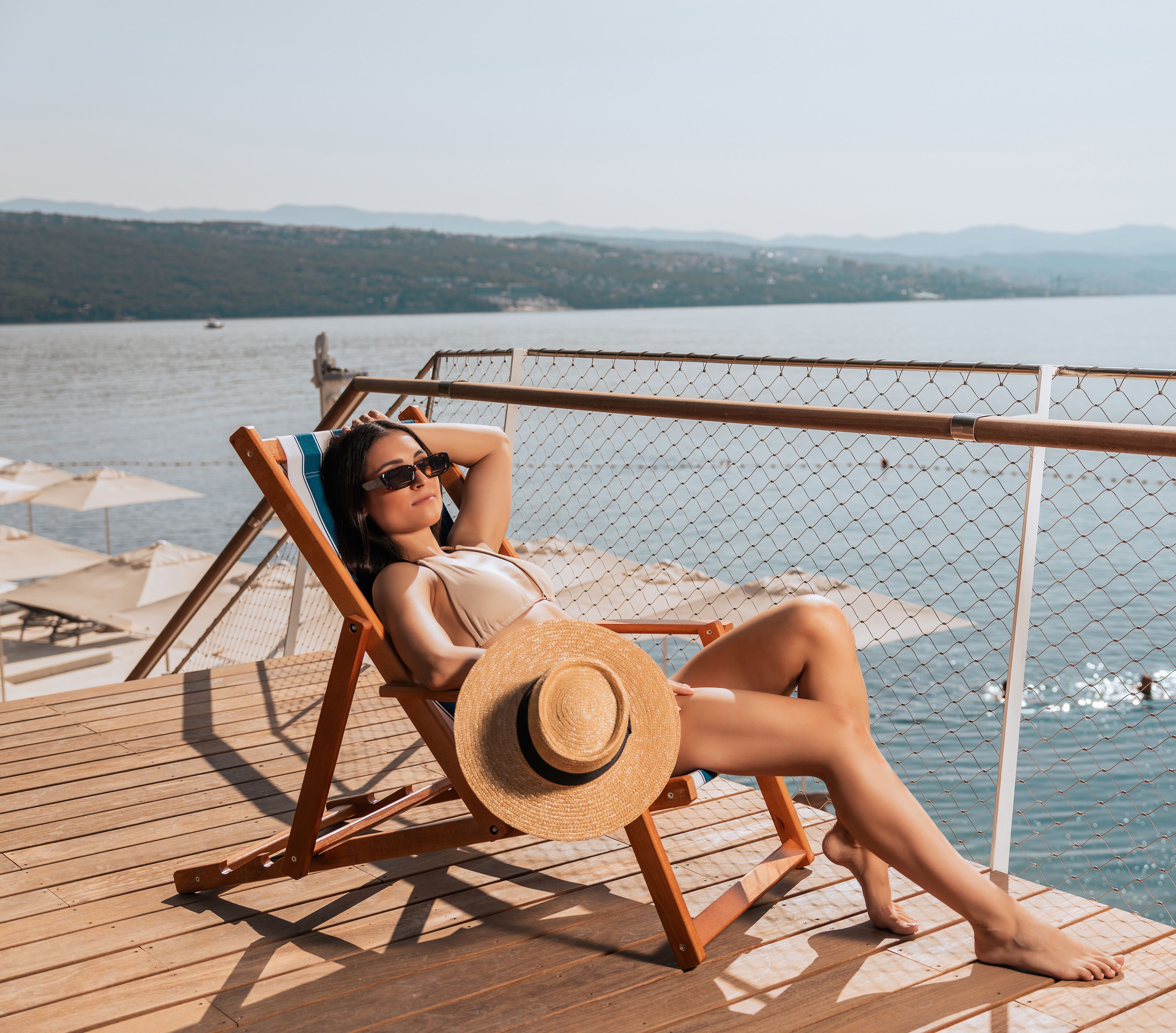 A person relaxing on a wooden deck chair with a straw hat, overlooking a body of water and distant mountains.
