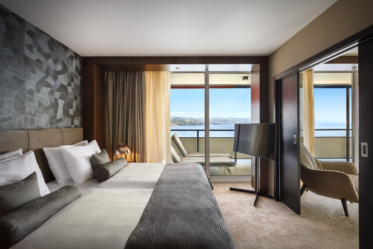 A modern hotel room with a large bed, a flat-screen TV, a balcony with a view of the sea, and a cityscape.
