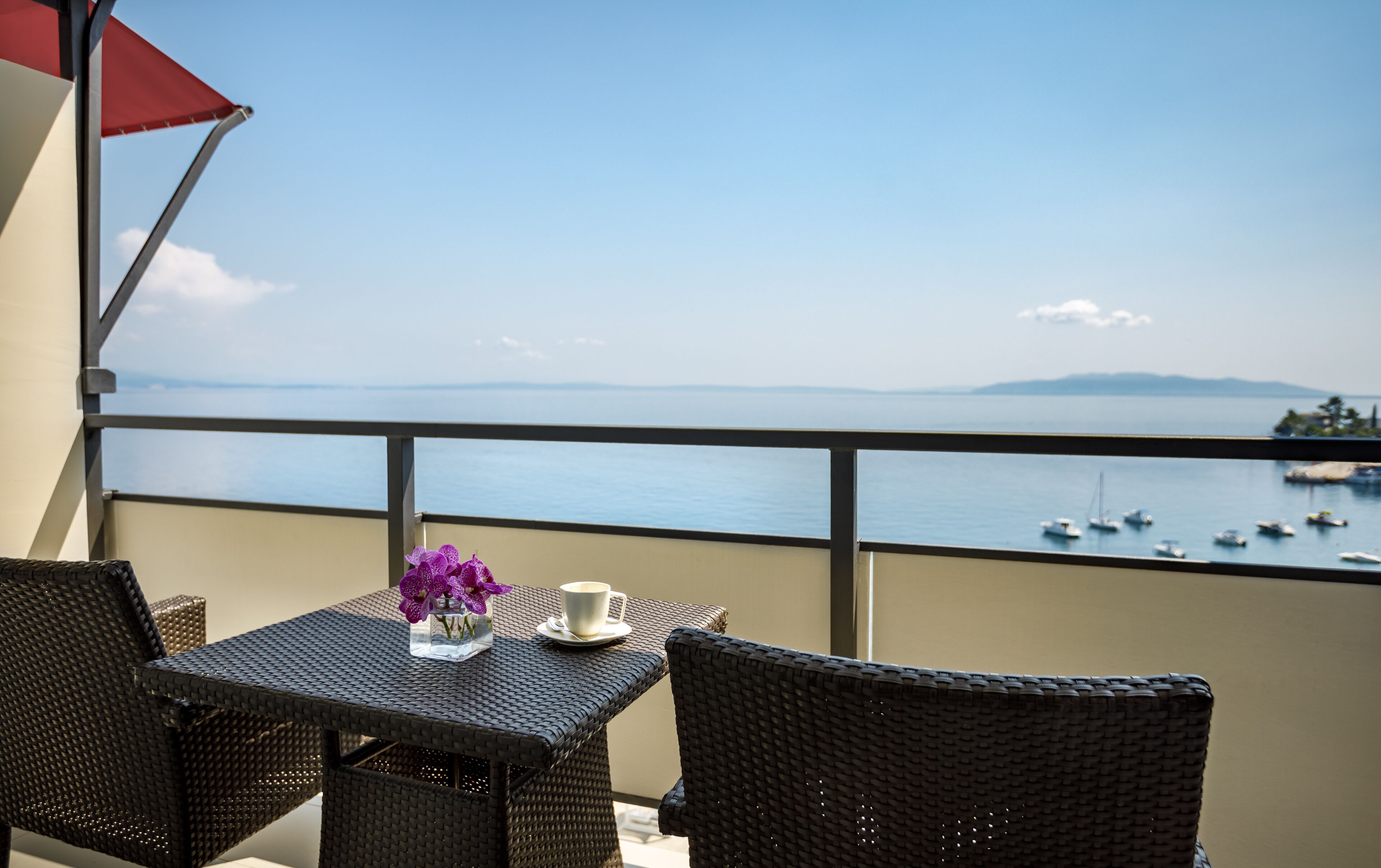 A balcony with wicker furniture, a table with a vase of purple flowers, and a view of the sea with boats.
