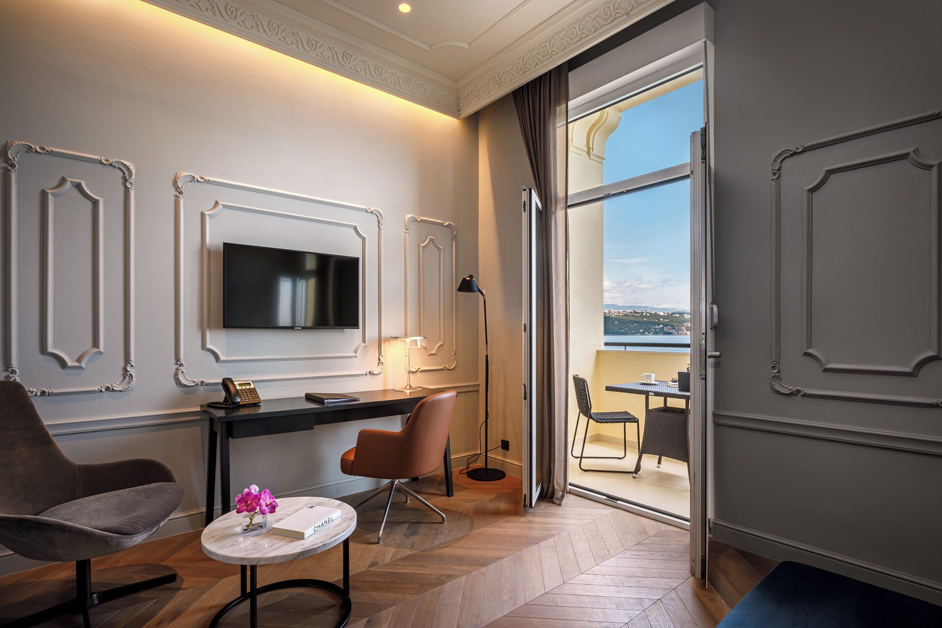 A modern hotel room with a large TV, a desk with a chair, a small round table with flowers, and a view of a body of water through a sliding door.