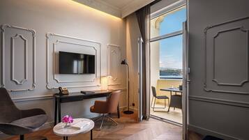 A modern hotel room with a large TV, a desk with a chair, a small round table with flowers, and a view of a body of water through a sliding door.