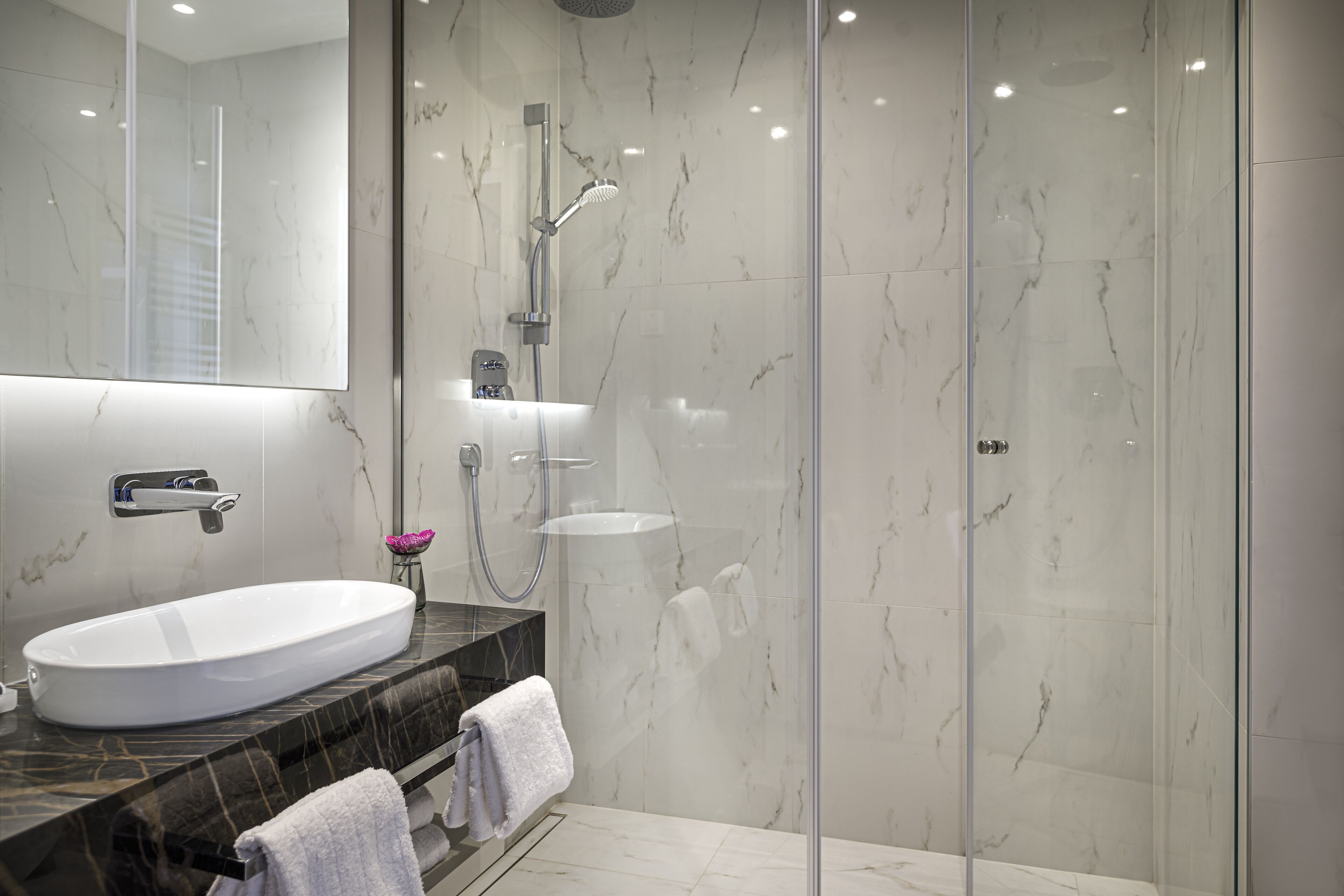 A modern bathroom with a large oval sink, a glass-enclosed shower, and a marble-tiled wall.