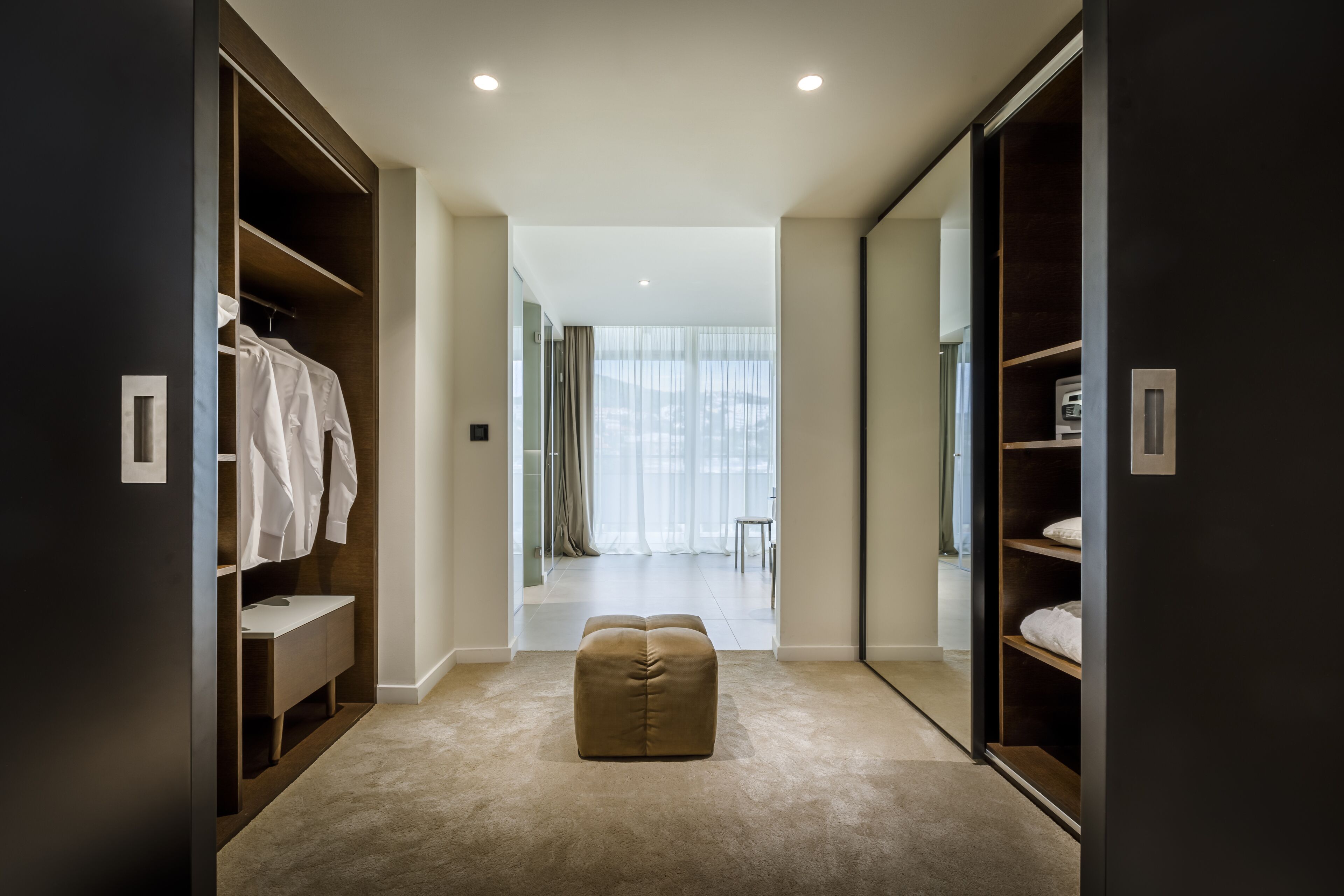 A modern walk-in closet with ample storage, a bench, and a view into a bright room.
