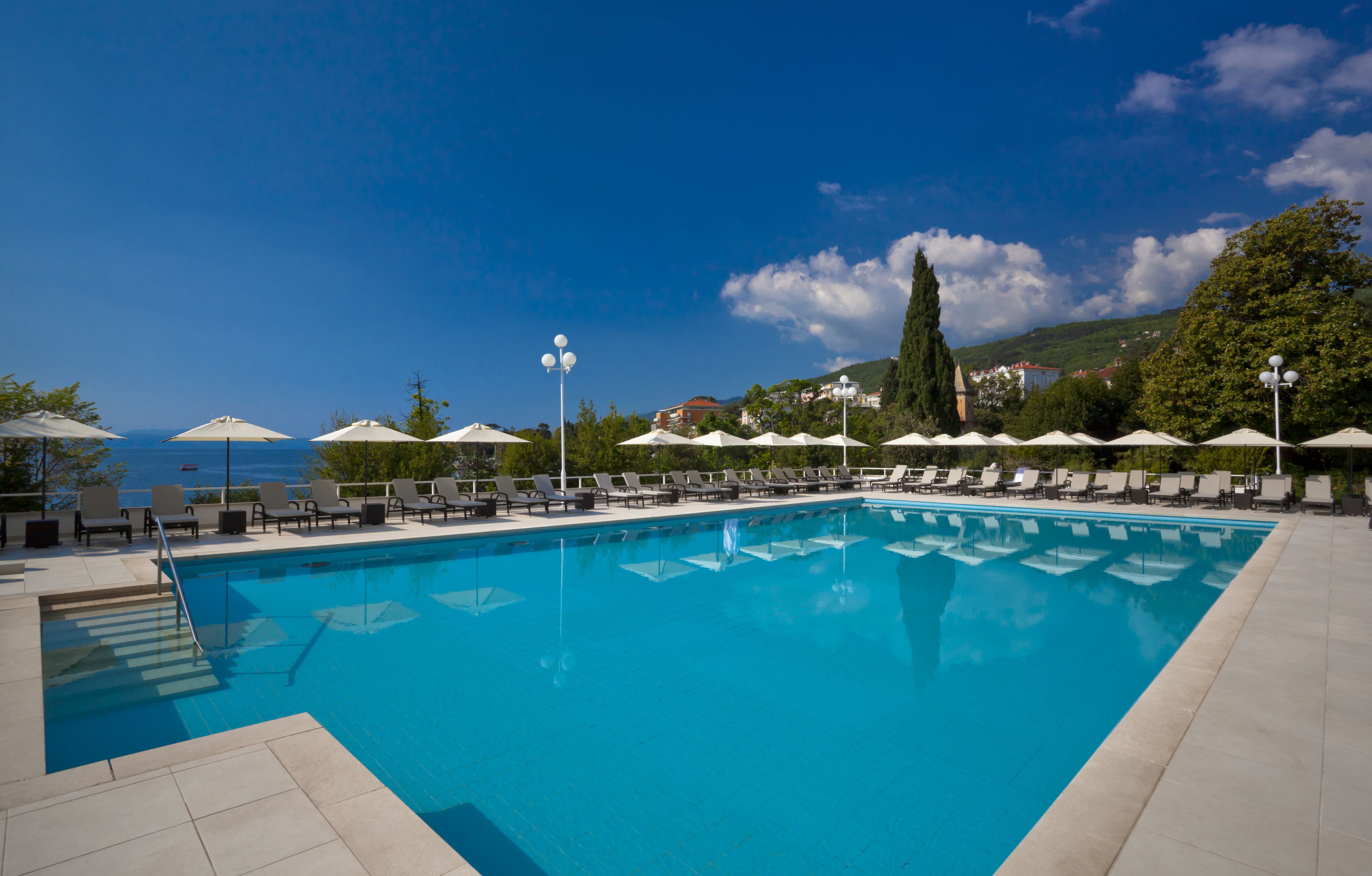 A rectangular swimming pool with clear blue water, surrounded by lounge chairs and umbrellas, with a view of the sea and hills in the background.