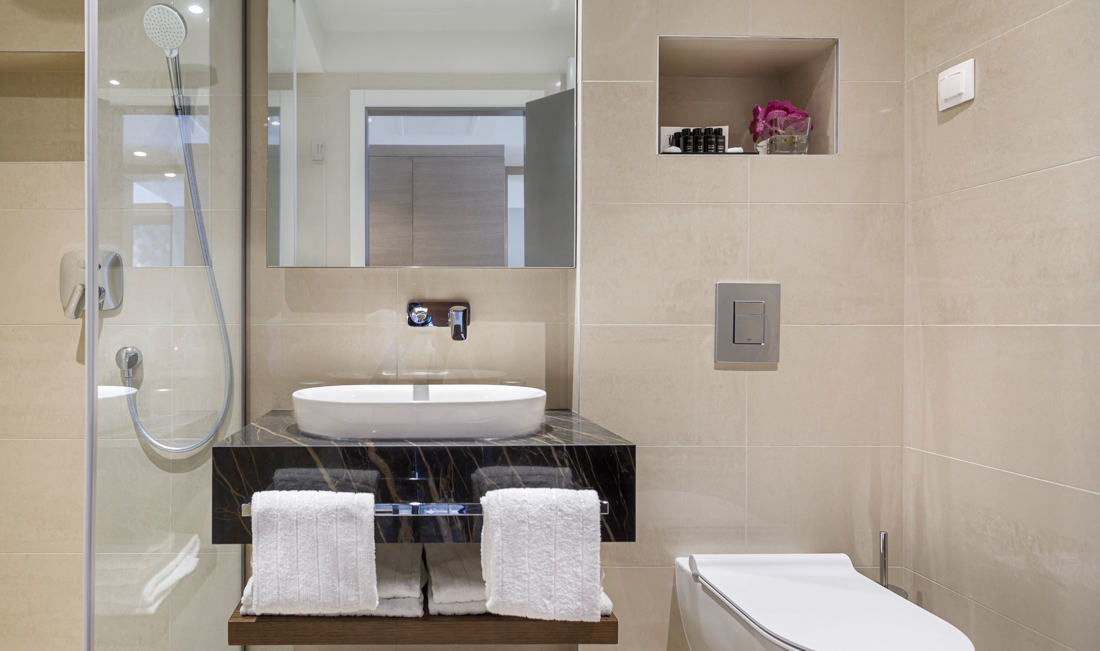A modern bathroom with a glass-enclosed shower, a marble countertop, a white sink, and a built-in shelf with toiletries.