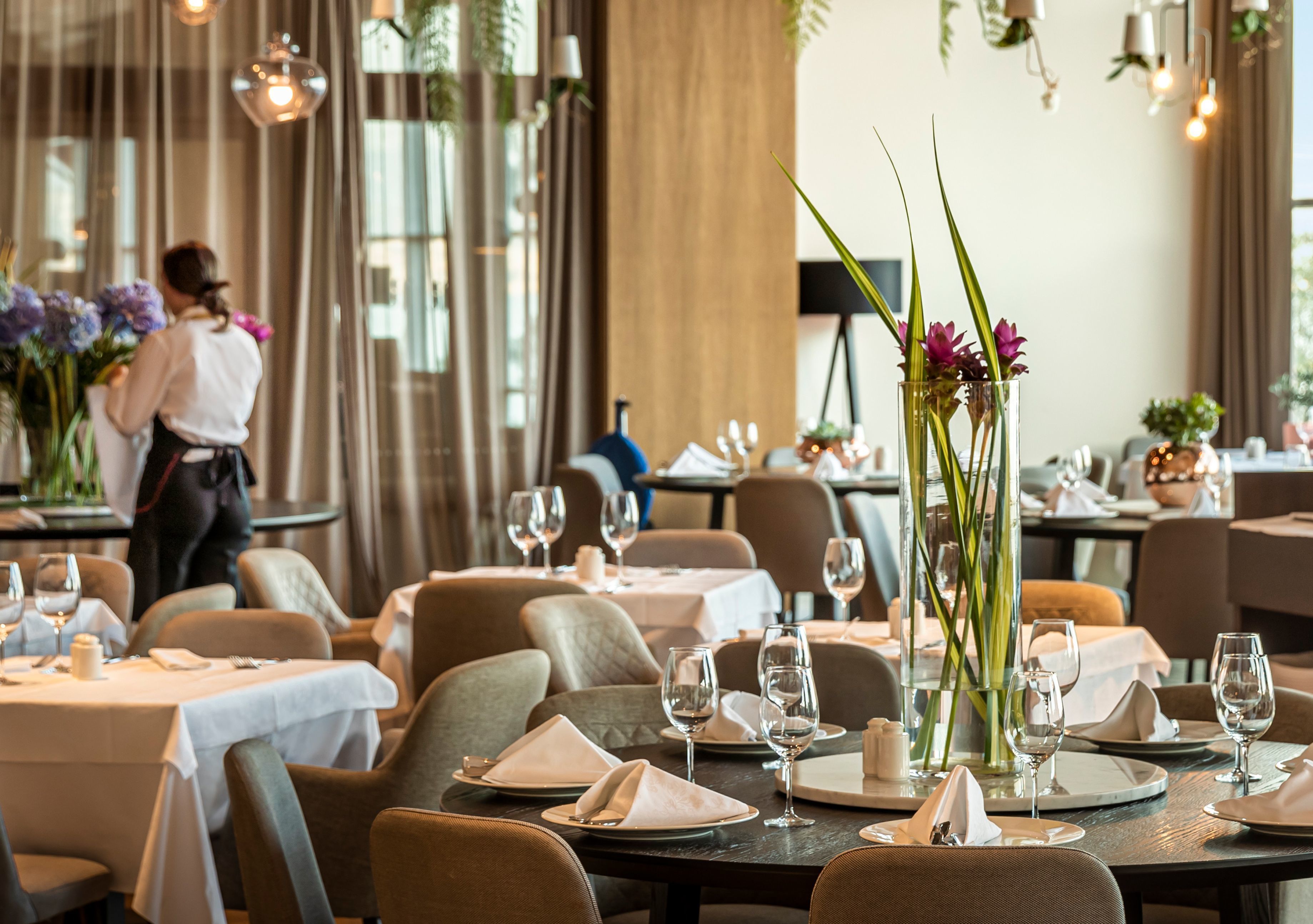 En restaurantscene med dækkede borde, en tjener i baggrunden og en vase med blomster på bordet.