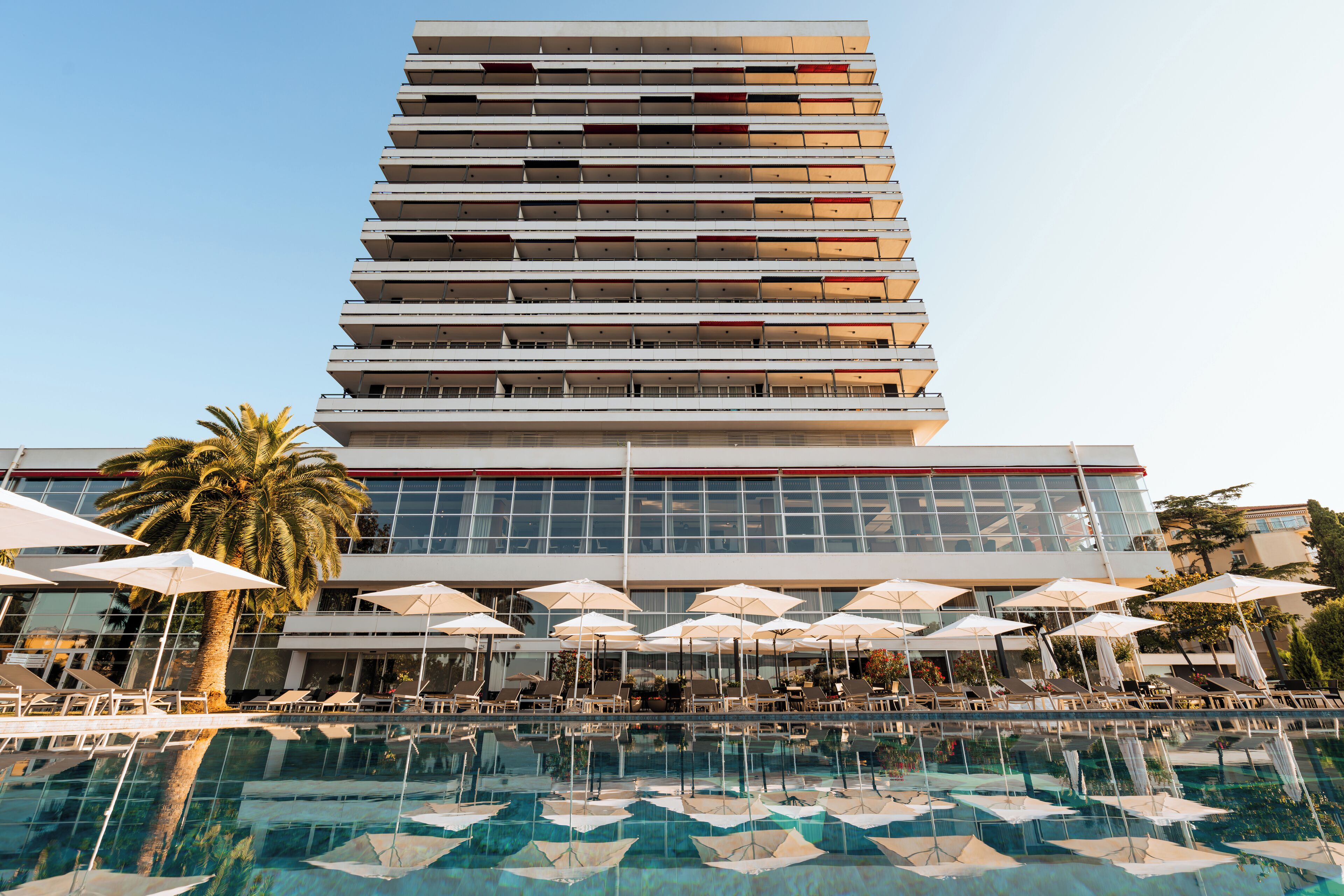 A modern hotel with a large pool area, multiple balconies, and outdoor umbrellas.