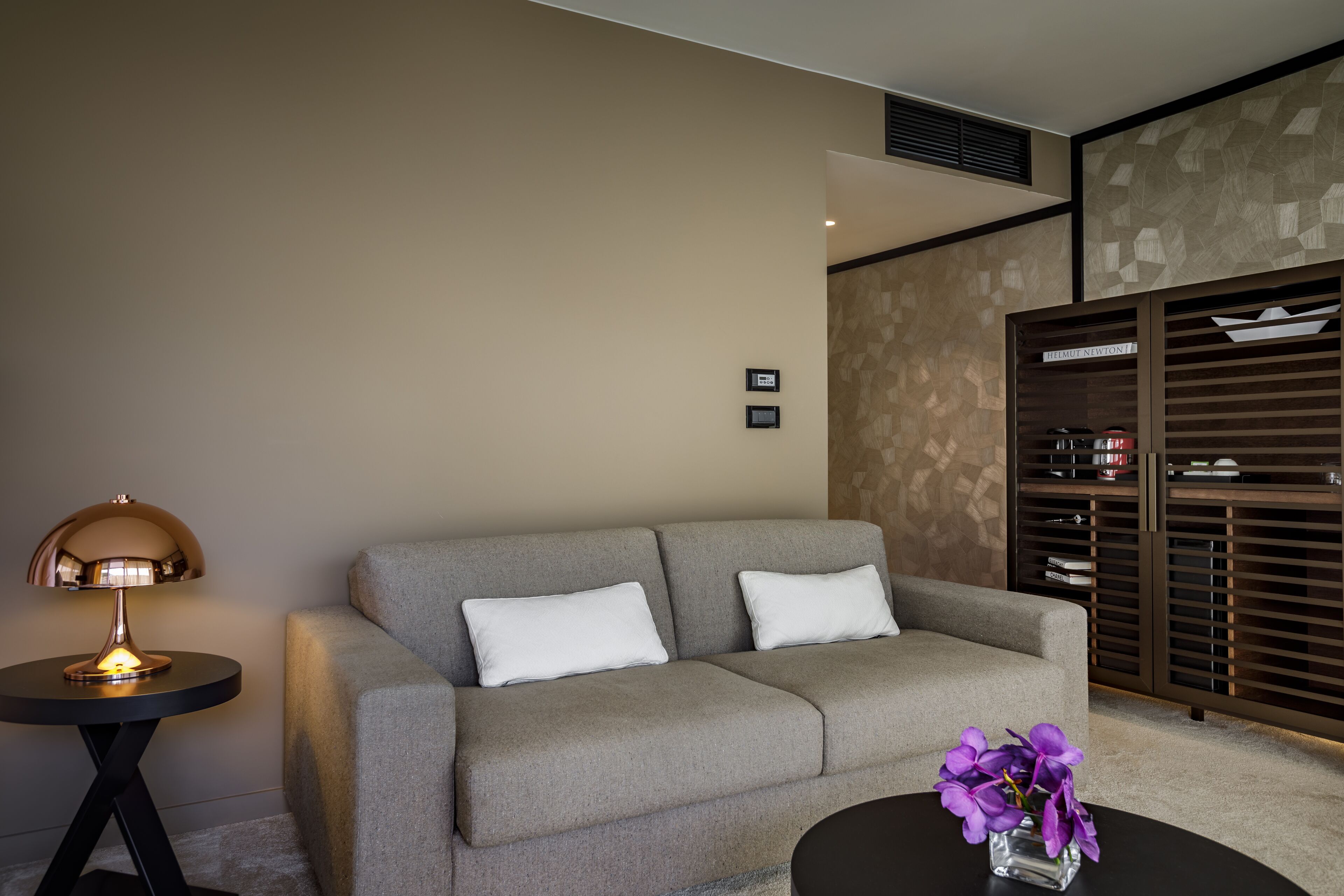 A modern living room with a grey sofa, a small round table with a vase of purple flowers, and a lamp on a side table.