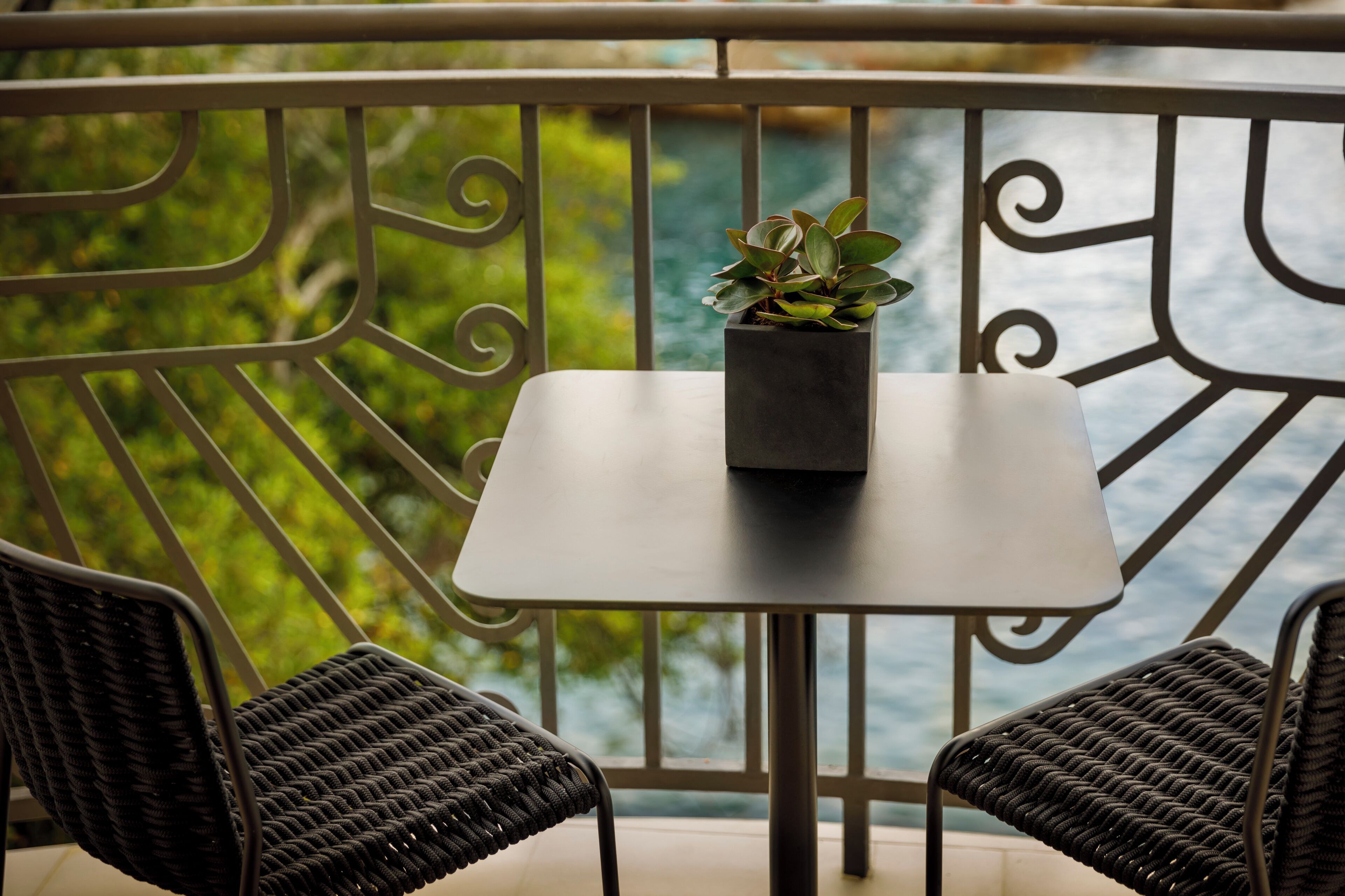 Un balcón con una mesa y dos sillas de mimbre, una planta en maceta sobre la mesa, y vistas a zonas verdes y al agua.