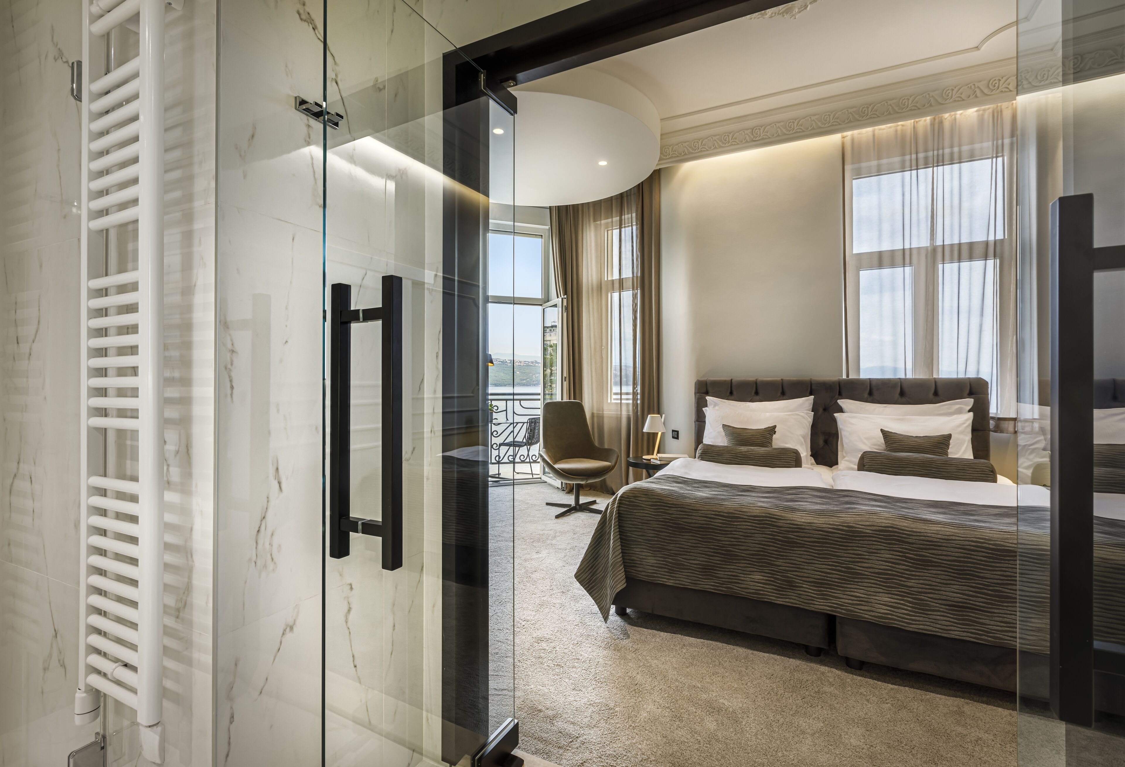 A modern hotel room with a large bed, a sitting area with a chair, and a view of the city through large windows.