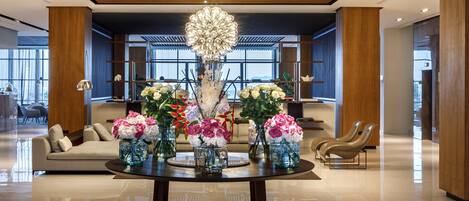 A modern lobby with a round table adorned with floral arrangements, a large chandelier, and a seating area with sofas and chairs.
